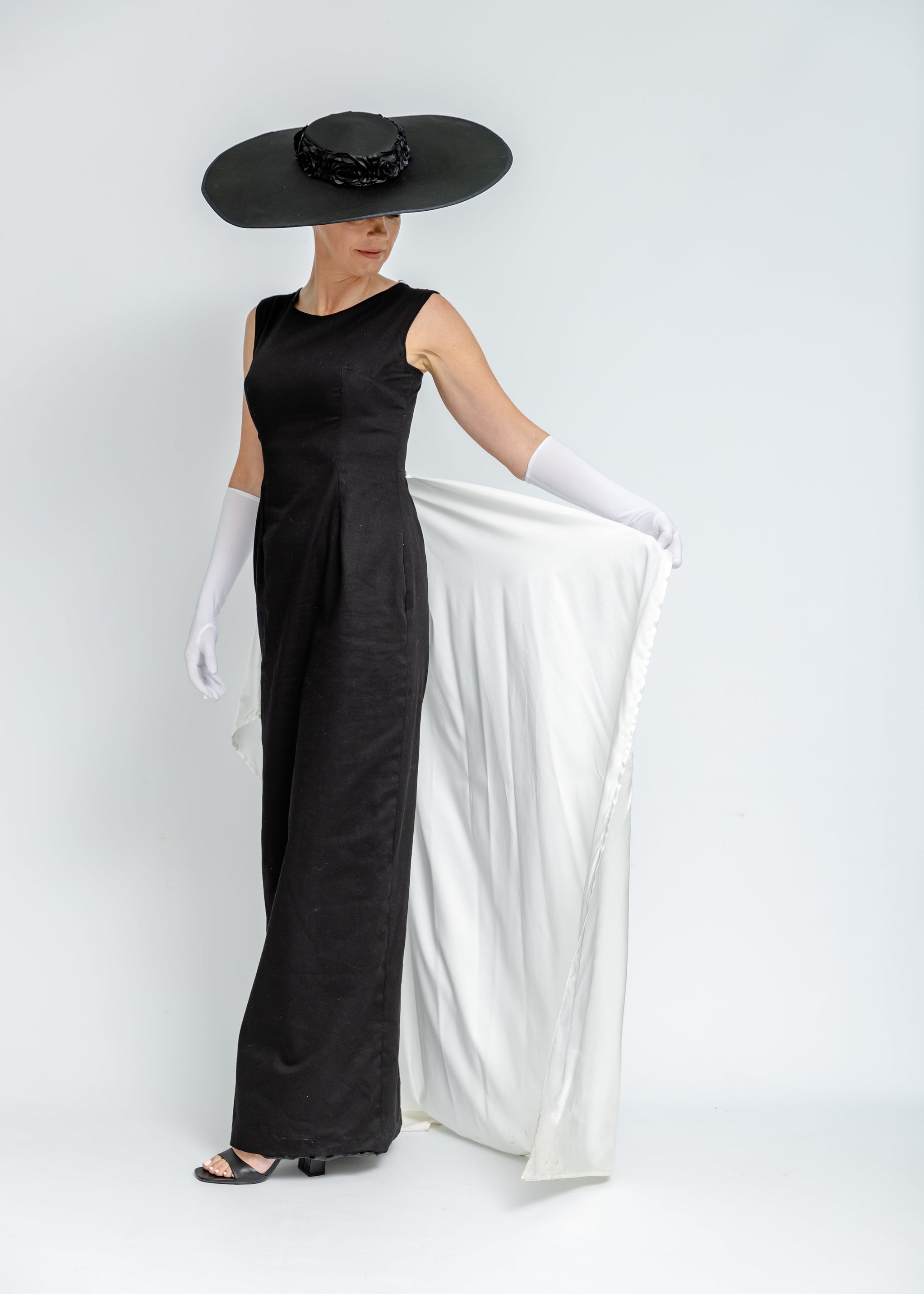 Person wearing a black dress, white gloves, and a large black hat with a white scarf.