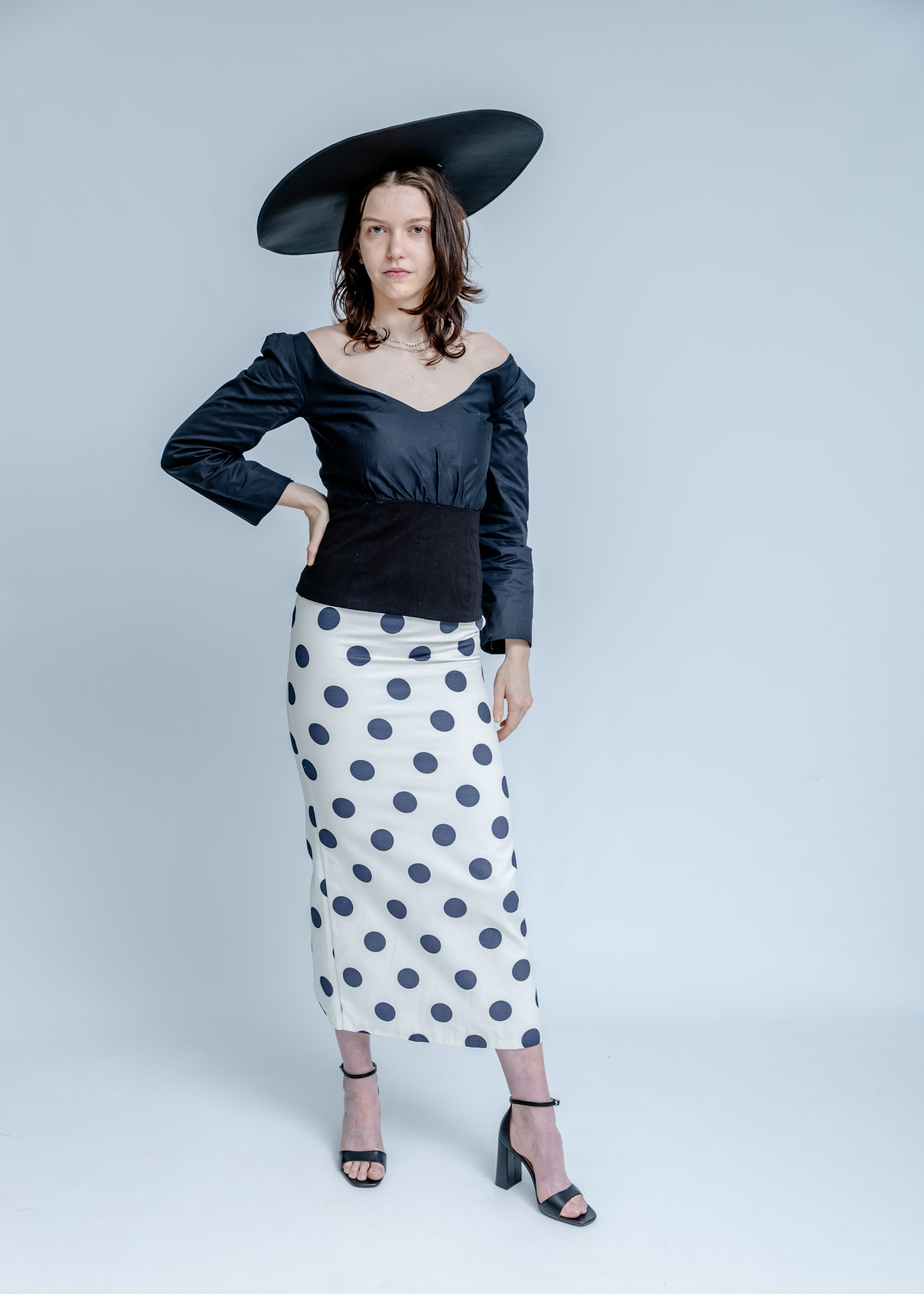 Woman wearing a black off-shoulder top and polka dot skirt with a wide-brimmed hat on a white background