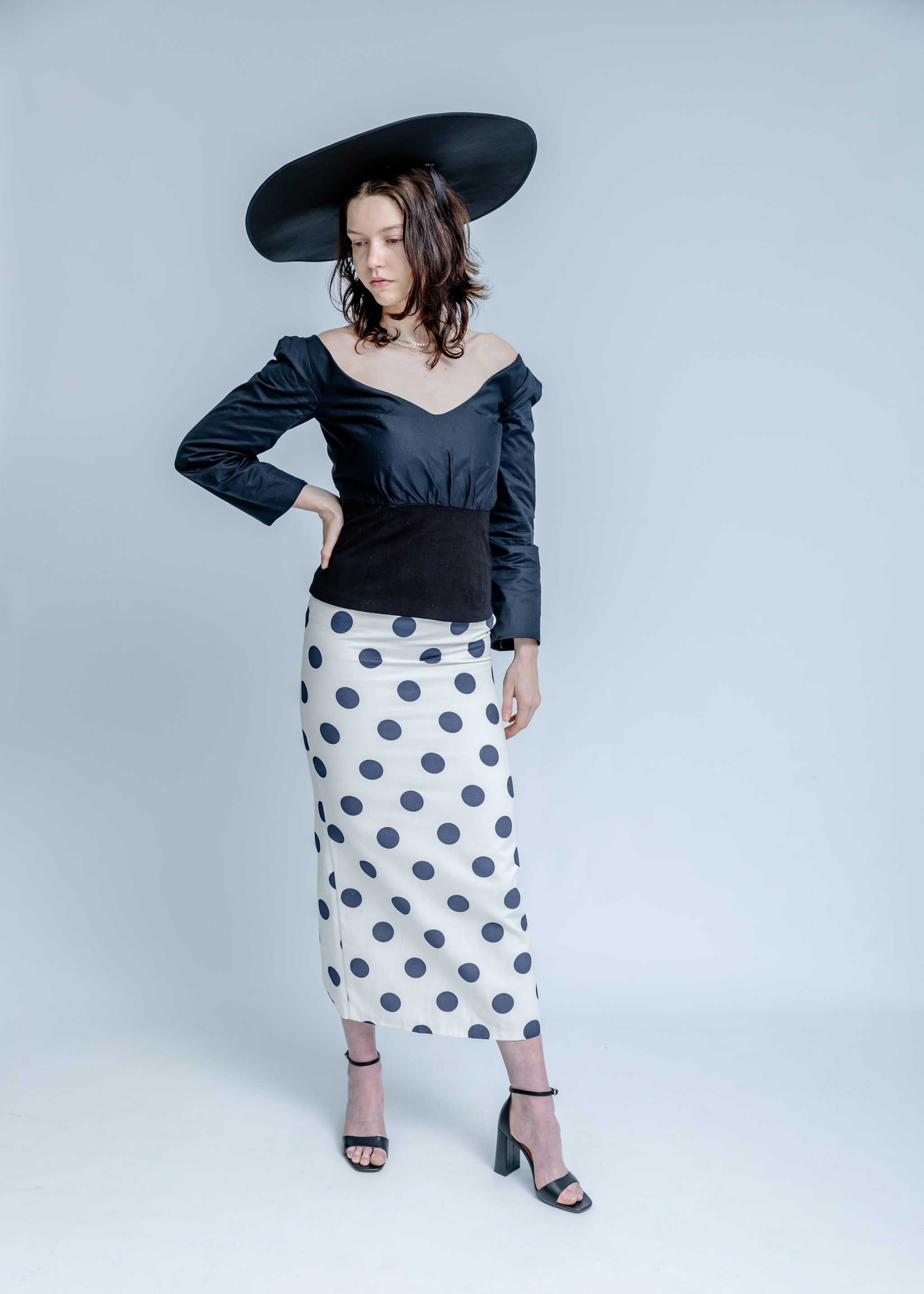 Woman wearing a black off-shoulder top and polka dot skirt with a wide-brimmed hat on a white background