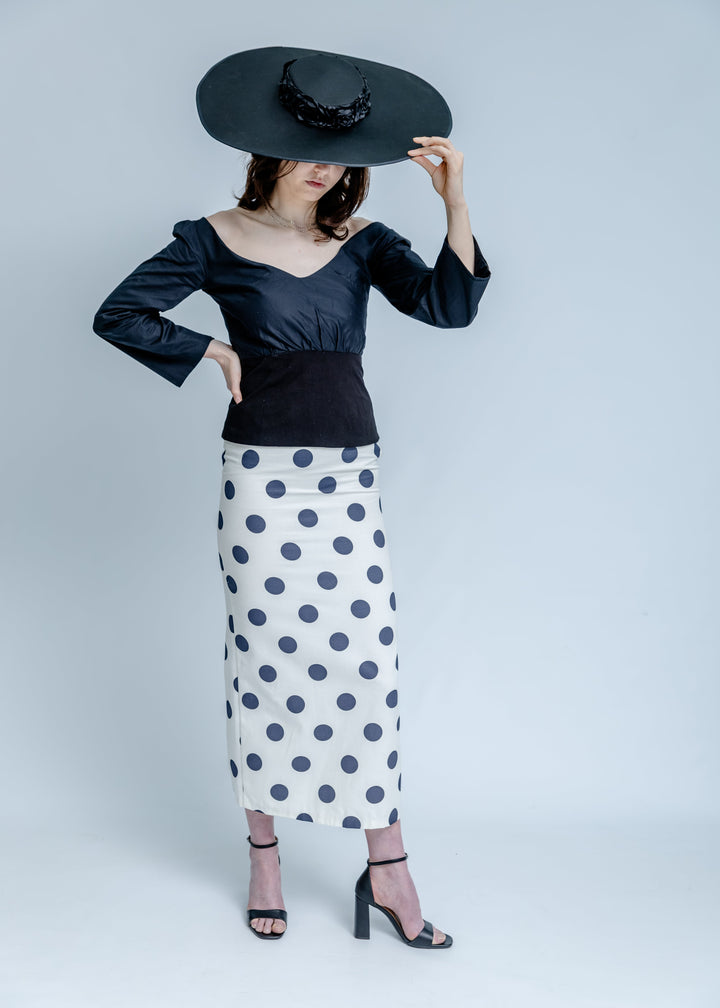 Woman wearing a black top and polka dot skirt with a wide-brimmed black hat on a white background