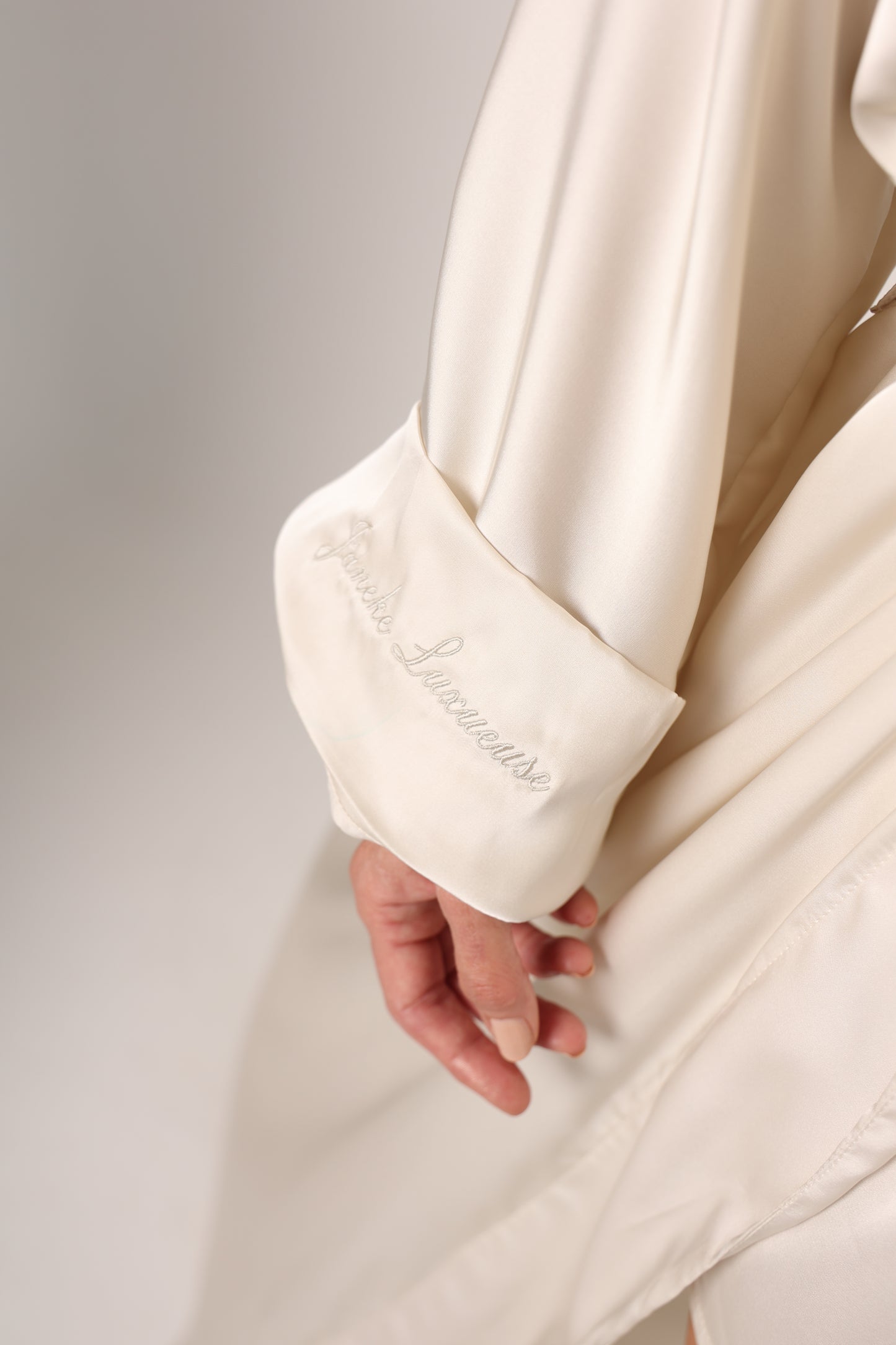 Close-up of a white satin robe with a hand holding it, on a light gray background.
