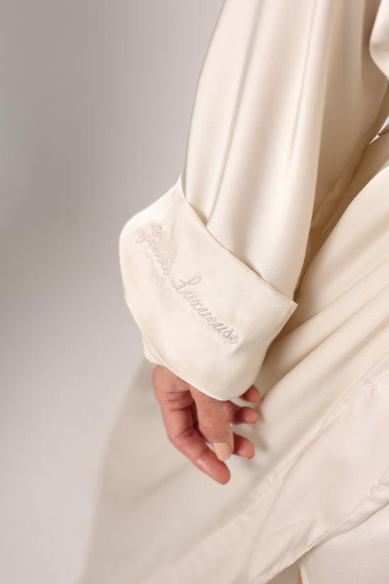 Close-up of a white satin robe with a hand holding it, on a light gray background.