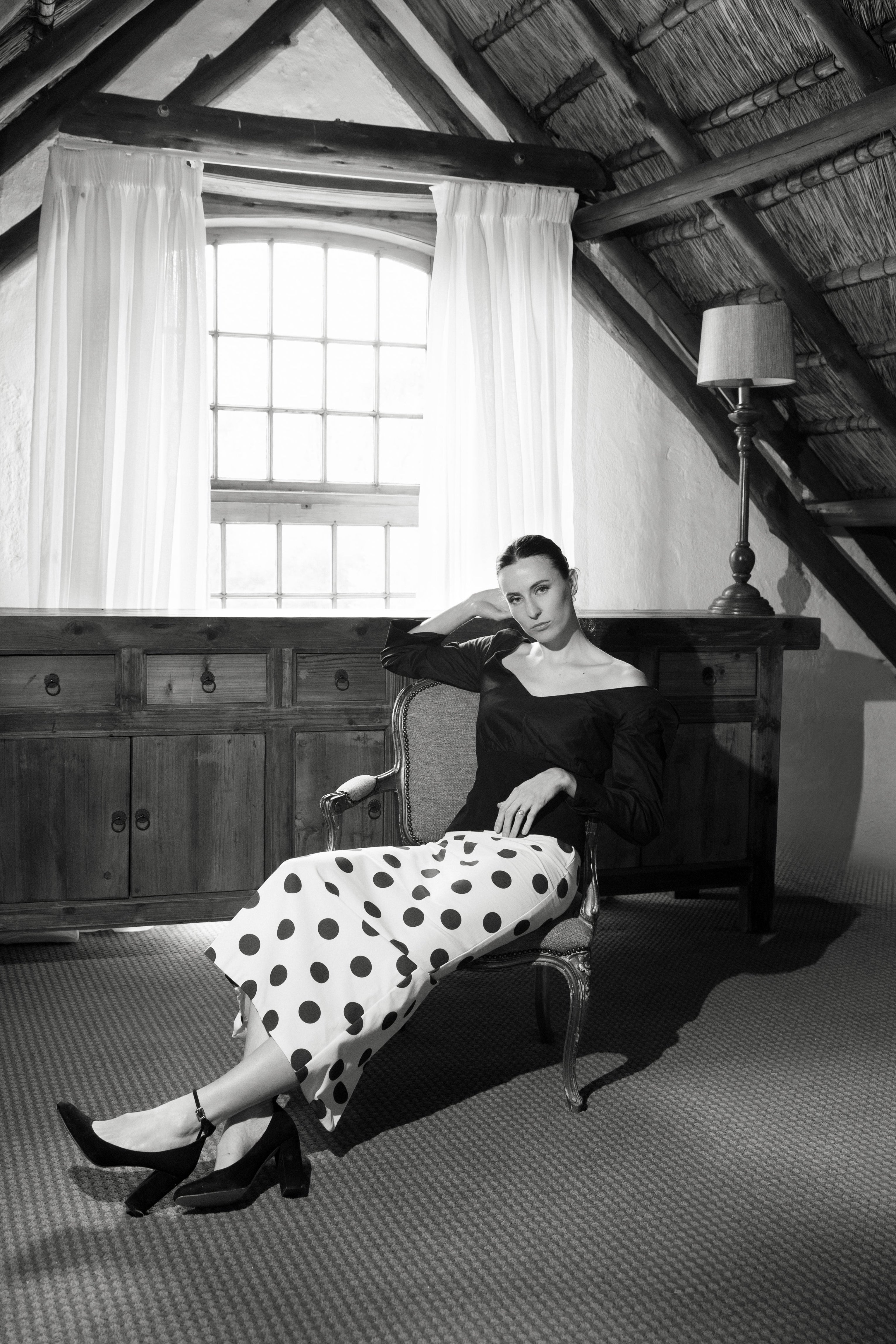 Woman in a polka dot dress sitting on an armchair in a room with wooden beams and a window.