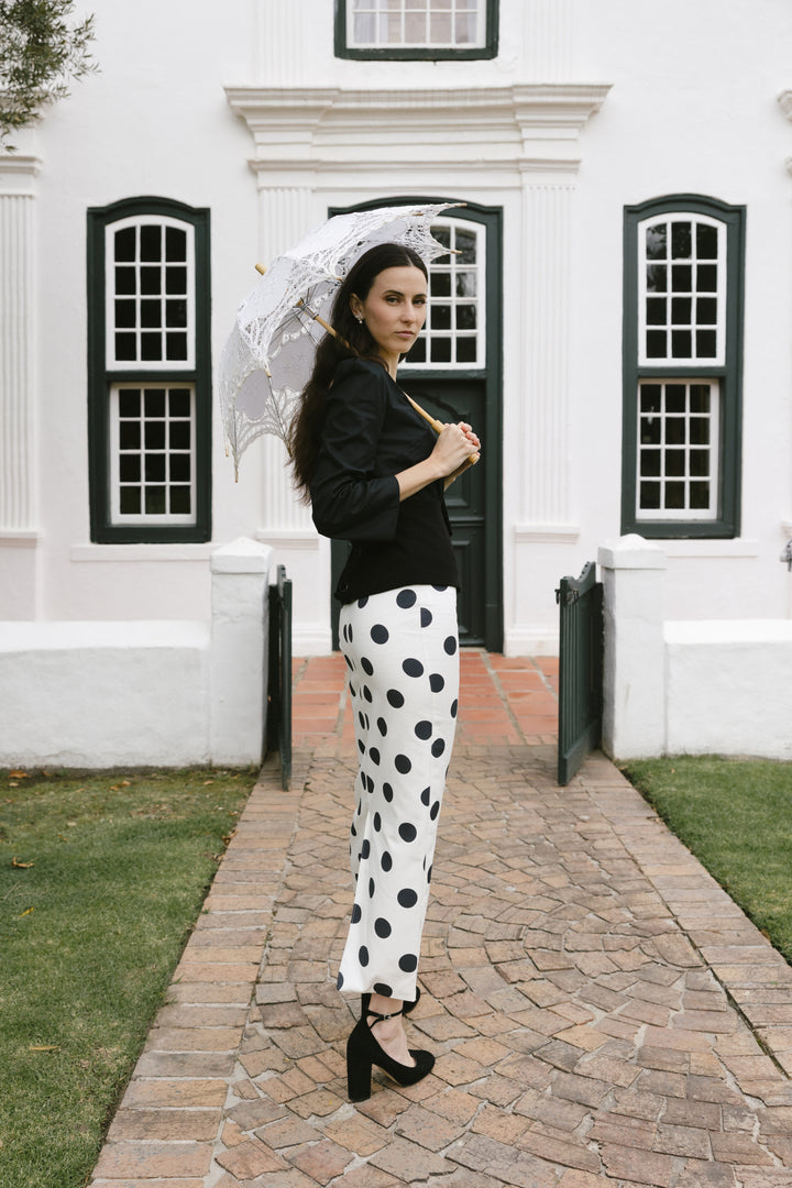 Woman in black top and white polka dot pants holding a white parasol in front of a white building with black trim.