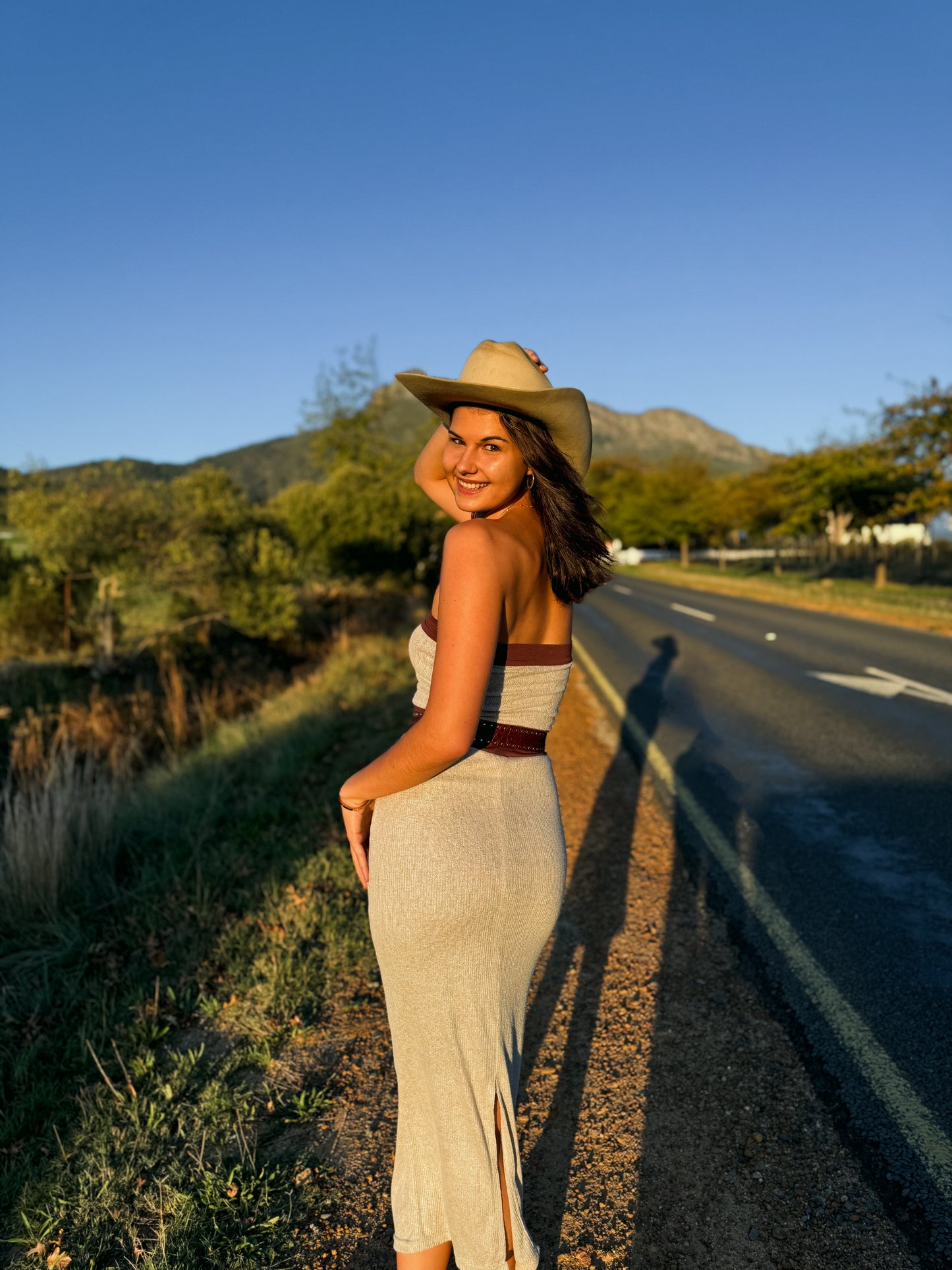 Woman in a dress and hat standing by a road with mountains in the background