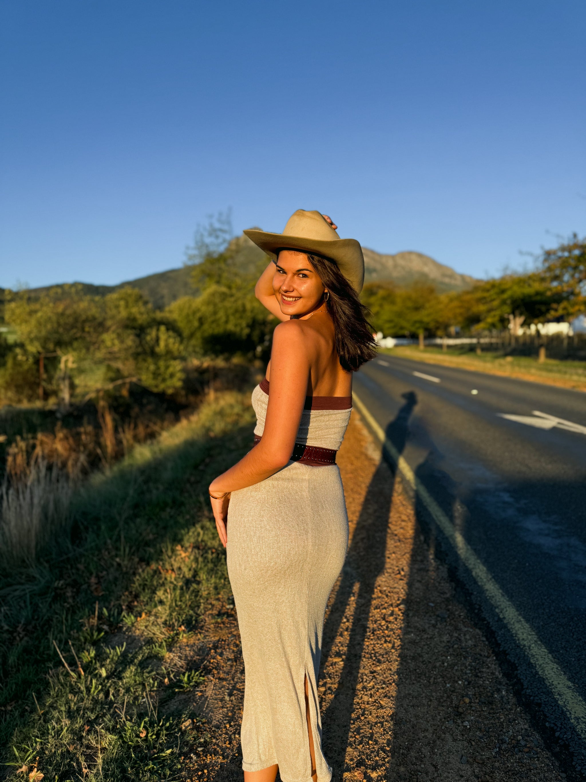 Woman in a dress and hat standing by a road with mountains in the background