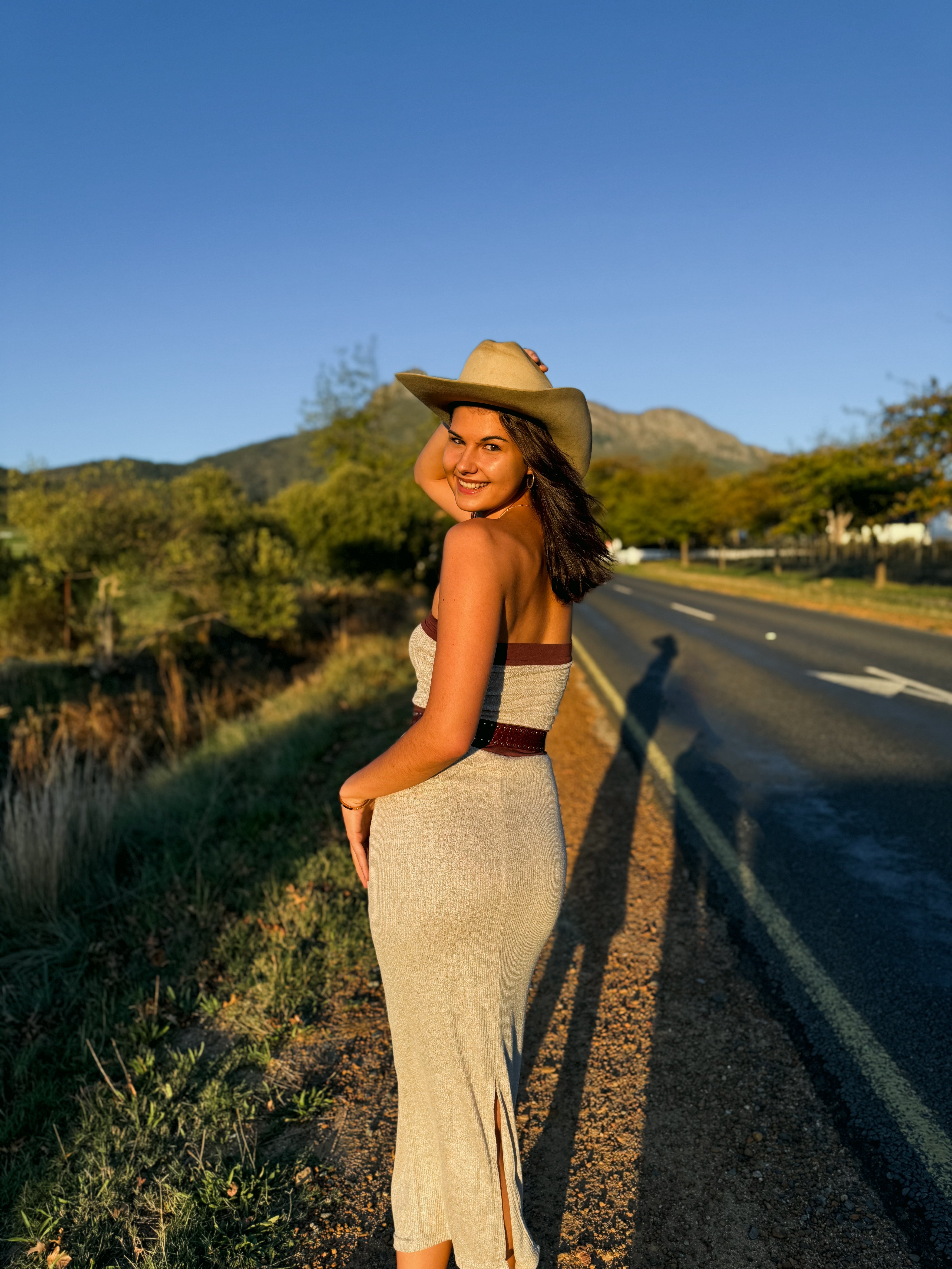 Woman in a dress and hat standing by a road with mountains in the background