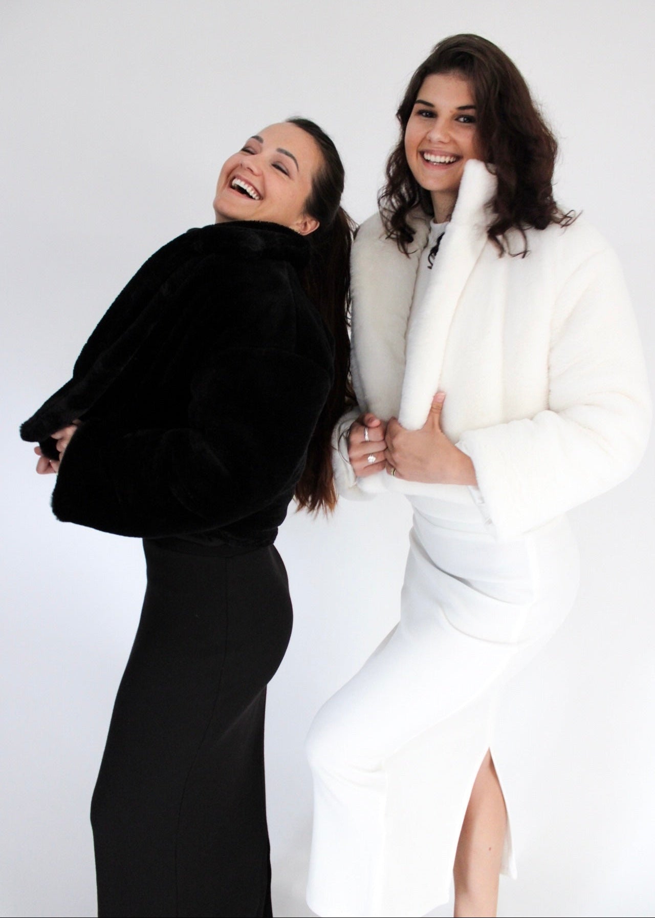 Two women, one in a black fur coat and the other in a white fur coat, posing against a white background.