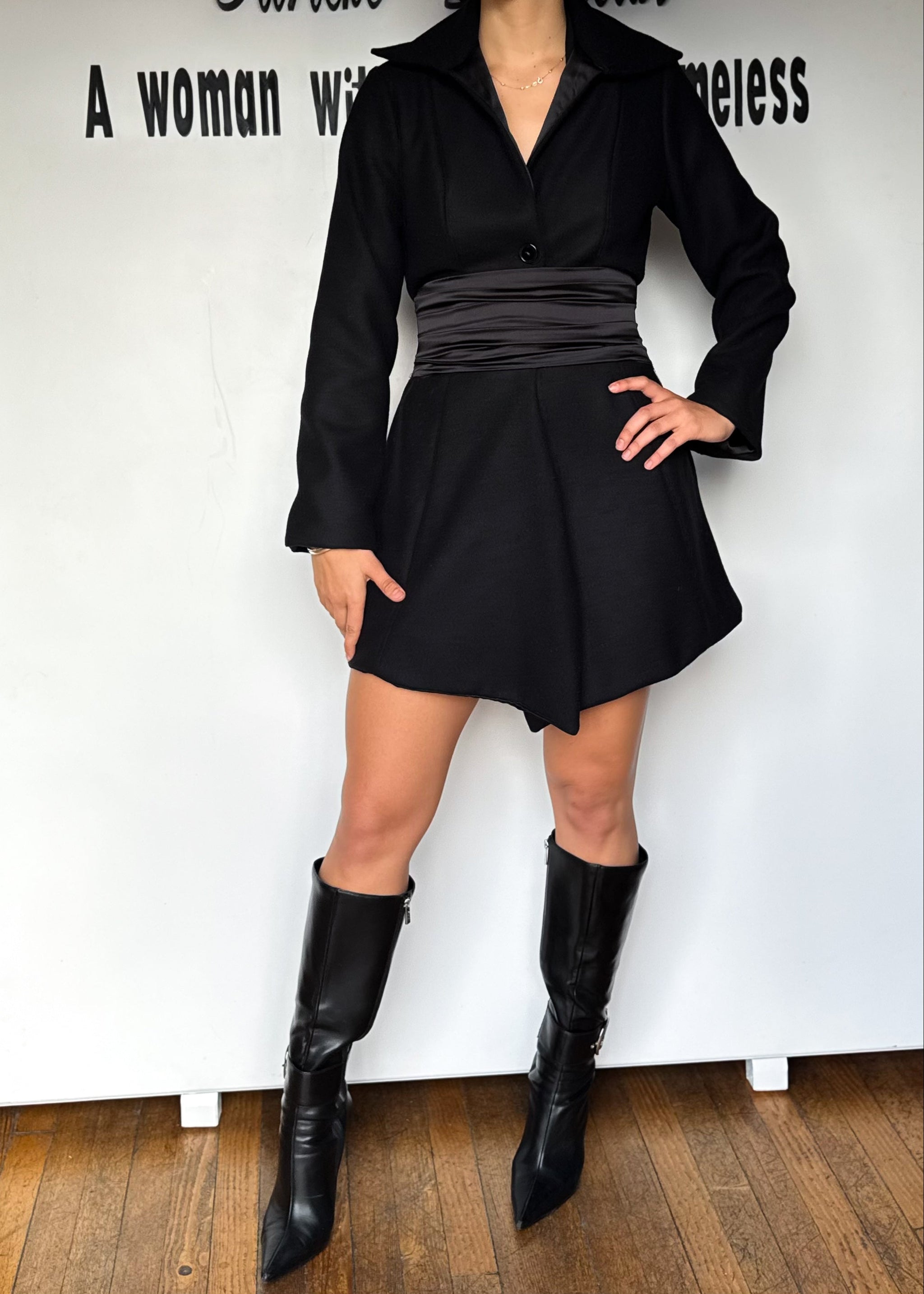 Person wearing a black outfit with a skirt and boots against a white wall.