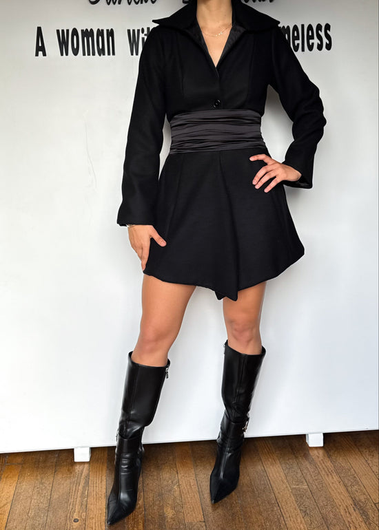 Person wearing a black outfit with a skirt and boots against a white wall.