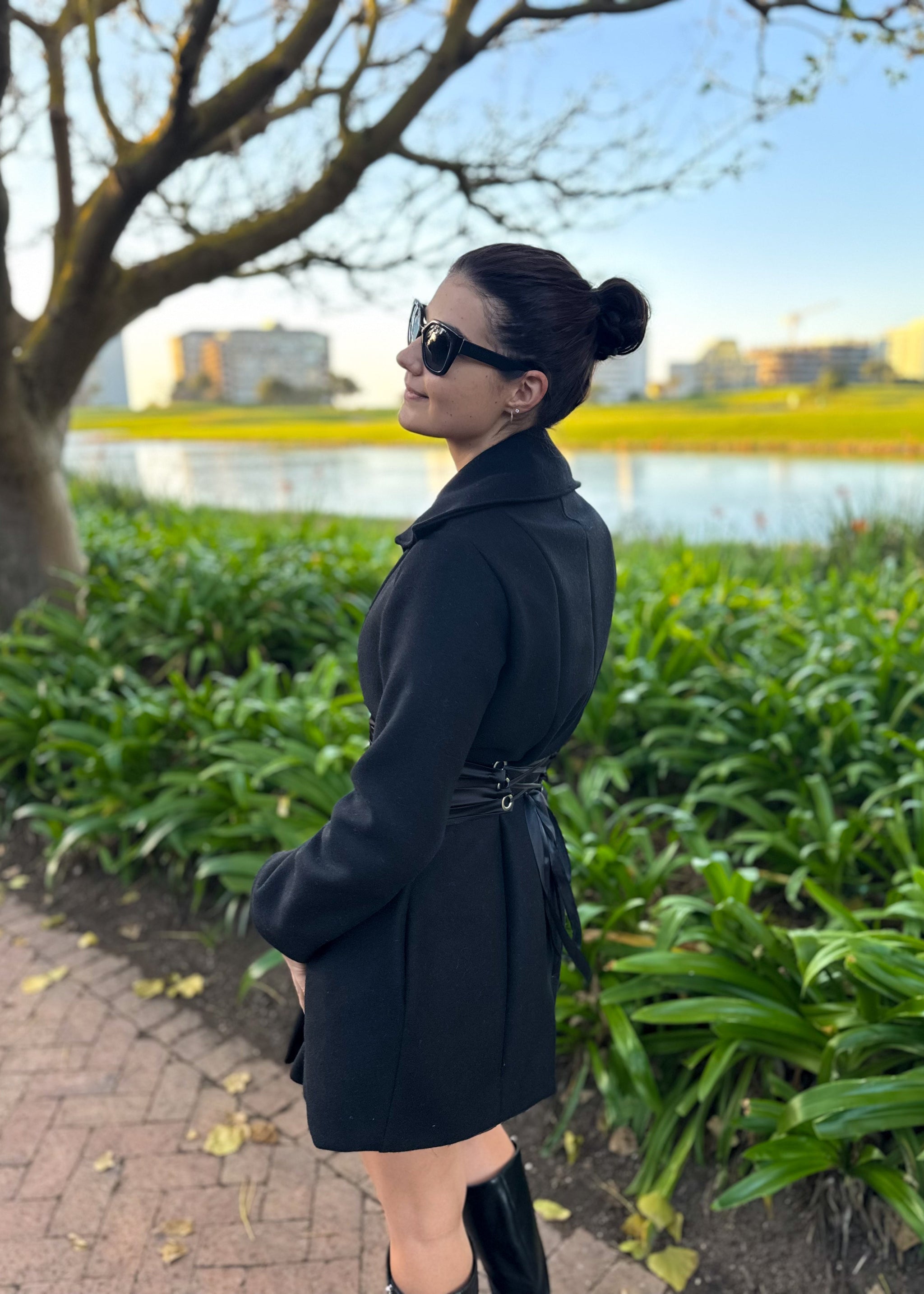 Woman in a black coat standing outdoors by a lake with greenery and buildings in the background.