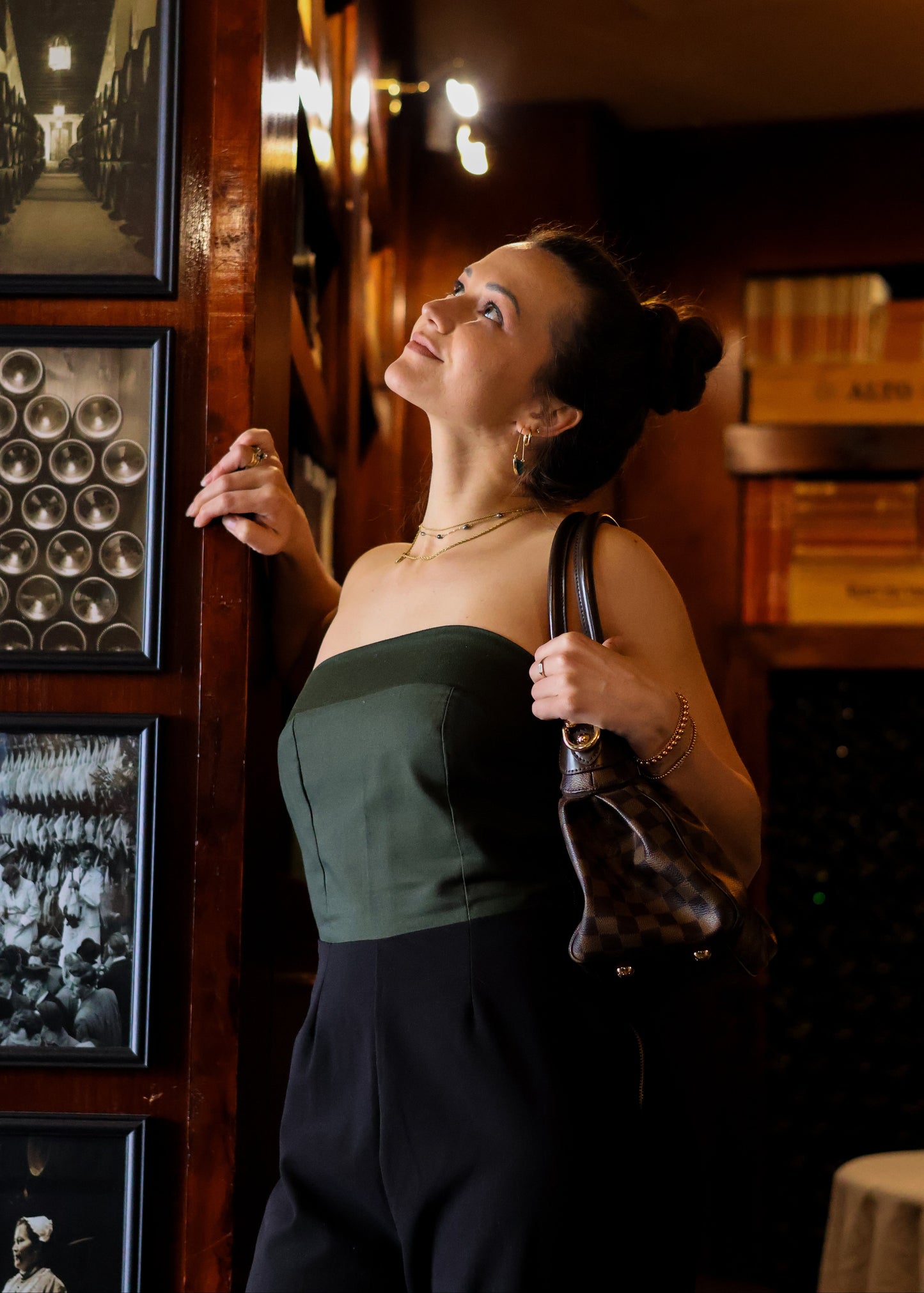 Woman in a green top and black pants standing in a dimly lit room with framed pictures on the wall.