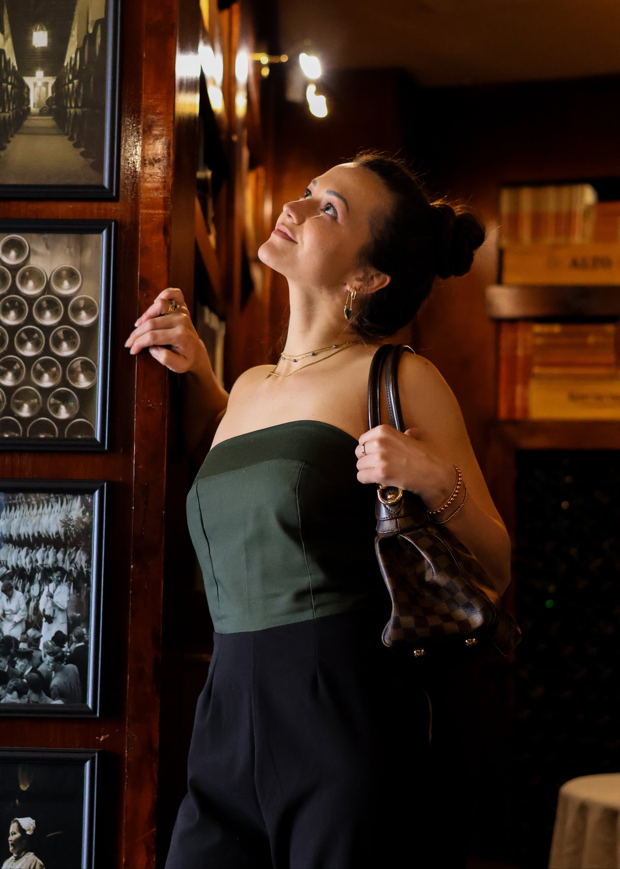 Woman in a green top and black pants standing in a dimly lit room with framed pictures on the wall.