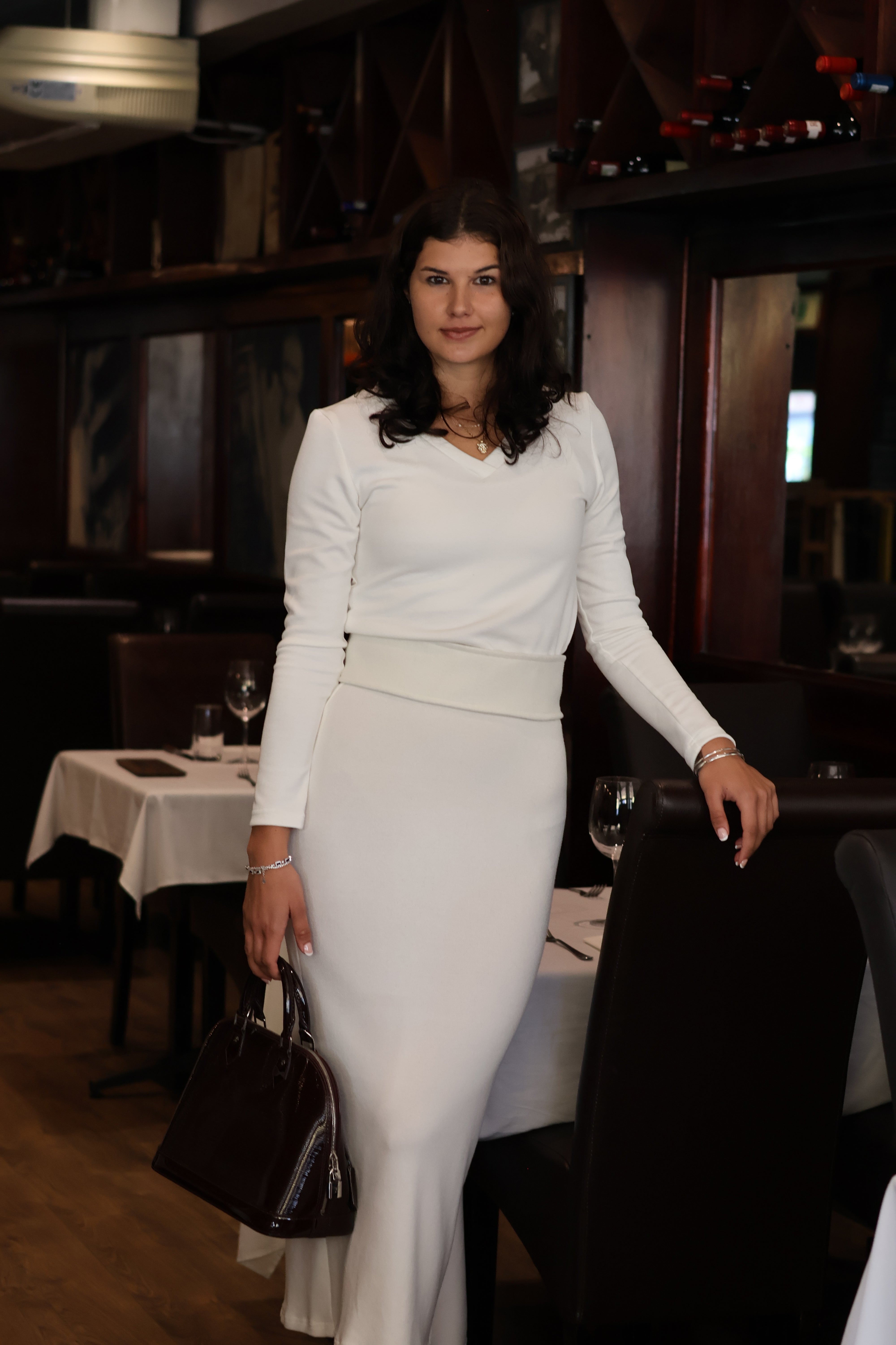 Woman in a white outfit standing in a dimly lit room with chairs and tables.