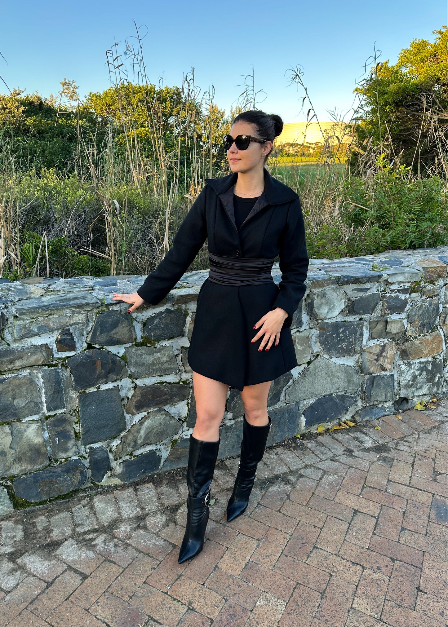 Woman in a black outfit standing by a stone wall with greenery in the background