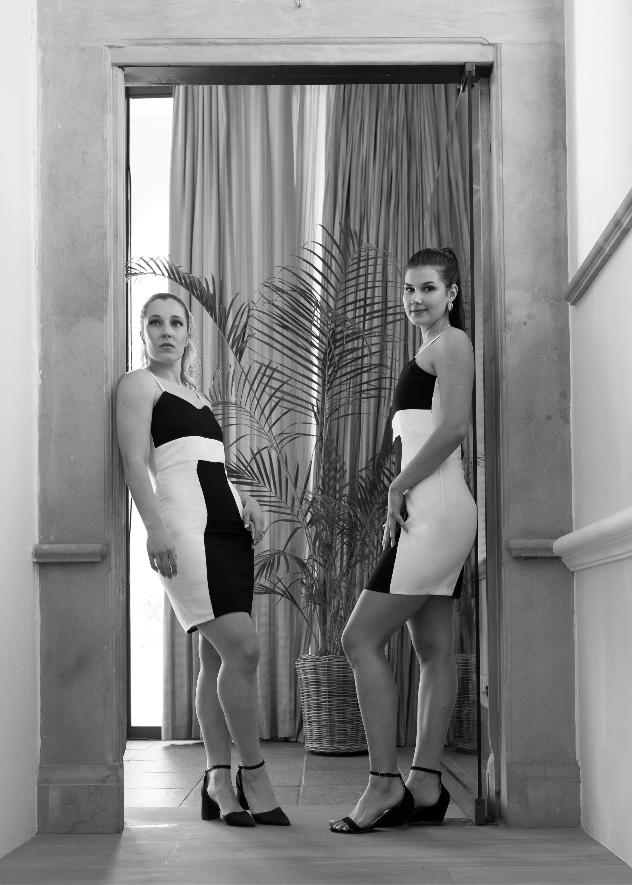 Two women in vintage-style outfits standing in a doorway with a plant and decorative elements.