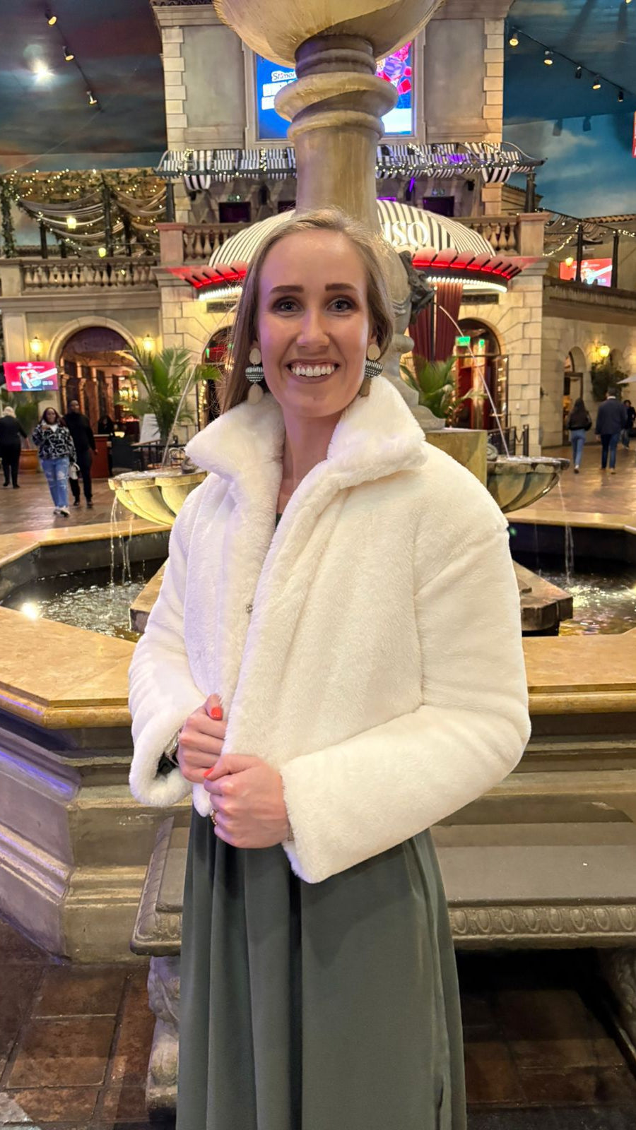Woman in a white coat standing in a decorative indoor area with fountains and people in the background.